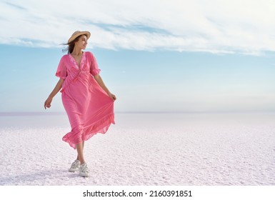 Adorable romantic woman admiring serenity colorful landscape of salt flats on pink lake. Girl wearing hat and flowing pink dress