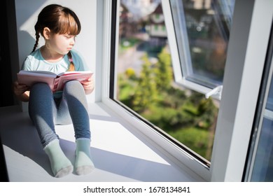 Adorable Sad Child Girl Reading By The Window. Quarantine. Coronavirus. Epidemic.