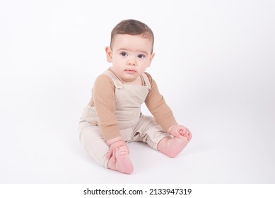 Adorable seven month old baby boy wearing beige overalls sitting on white background and looking to the camera front view. Childhood and innocence concept.  - Shutterstock ID 2133947319