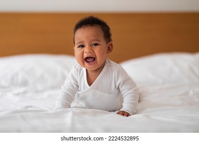 Adorable Small Black Baby Crawling On Bed And Laughing, Portrait Of Cute Little African American Infant Boy Or Girl Lying On His Tummy, Relaxing On White Bedsheets In Bedroom At Home, Copy Space