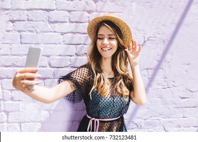 Adorable Smiling Girl In Trendy Straw Hat Making Selfie While Waiting Friend Outside. Outdoor Portrait Of Graceful Laughing Lady Taking Picture Of Herself On Old Wall Background.