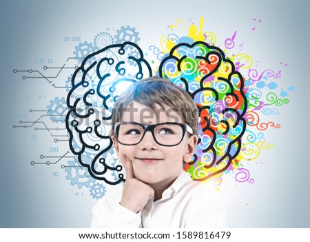 Adorable smiling little boy with glasses standing near gray wall with colorful left and right brain sketch. Concept of brainstorming and education.