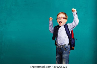 Adorable Smiling Little Kid With Big Backpack Jumping And Having Fun Against Blue Wall. Looking At Camera. School Concept. Back To School