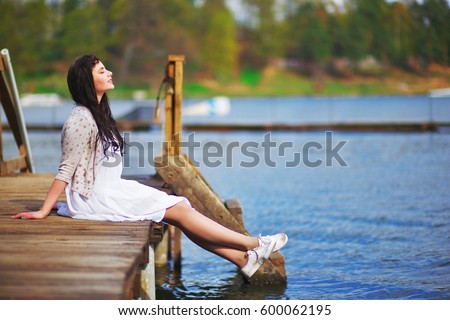 Adorable smiling long-haired brunette girl in a white dress and beige knitted cardigan sitting on wooden dock over the water with my eyes closed and enjoying warm spring Sunny day free life, side view
