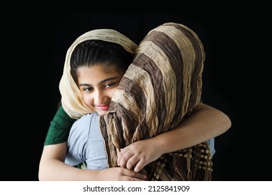 Adorable smiling Pakistani Muslim girl with beautiful eyes wearing hijab, hugging her mother on dark black background, warm love in the family of mom and daughter.