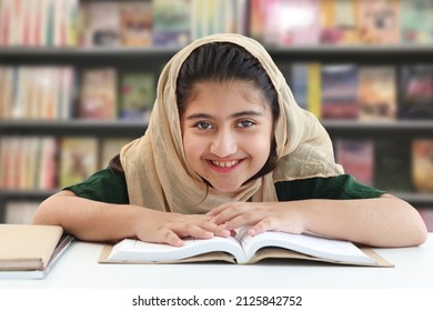 Adorable smiling Pakistani Muslim girl with beautiful eyes wearing hijab, studying and doing homework on table, happy student kid reading book on blurred background of bookcase in library. - Powered by Shutterstock