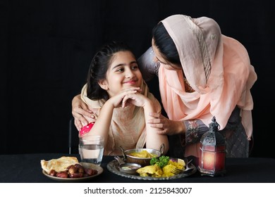 Adorable Smiling Pakistani Muslim Girl With Beautiful Eyes Sitting At Kitchen Table, Mother With Hijab Serving Islamic Halal Food On Dark Black Background, Mom Take Care Daughter. Family Warmth Love.