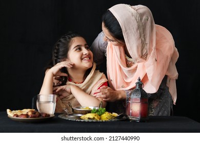 Adorable Smiling Pakistani Muslim Girl With Beautiful Eyes Sitting At Kitchen Table, Mother With Hijab Serving Islamic Halal Food On Dark Black Background, Mom Take Care Daughter. Family Warmth Love.