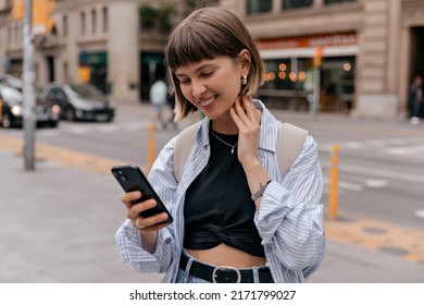 Adorable smiling straight short hair smiling while holding smartphone in the city wearing blue shirt and black top. Outside phot of caucasian woman wallking in city in warm day with backpack