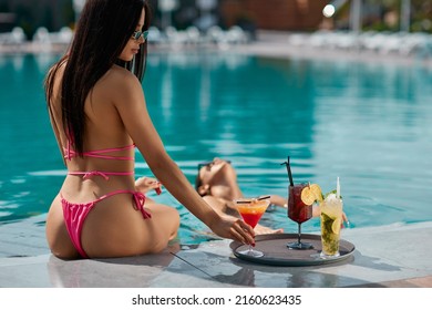 Adorable Tanned Lady In Pink Bikini Choosing Cocktail, While Sitting On Pool Edge At Resort. Back View Of Sexy Female In Thong Drinking Tasty Beverage, While Resting At Poolside. Concept Of Pleasure.