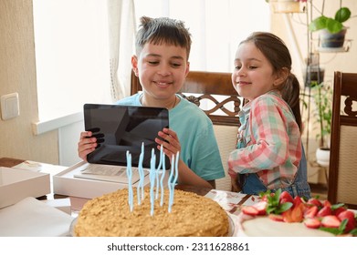 Adorable teen boy smiles rejoicing at a digital tablet for his birthday, sitting at table with delicious homemade cake, celebrating his birthday. People. Lifestyle. Technology. Life events. Childhood - Shutterstock ID 2311628861