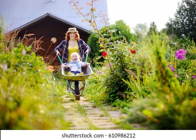 Adorable Toddler Boy Having Fun In A Wheelbarrow Pushing By Mum In Domestic Garden, On Warm Sunny Day. Active Outdoors Games For Kids In Summer.