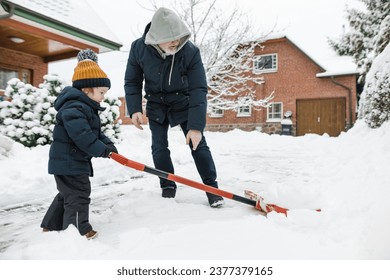 Adorable toddler boy helping his grandfather to shovel snow in a backyard on winter day. Cute child wearing warm clothes playing in a snow. Winter activities for family with kids.