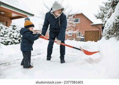 Adorable toddler boy helping his grandfather to shovel snow in a backyard on winter day. Cute child wearing warm clothes playing in a snow. Winter activities for family with kids.