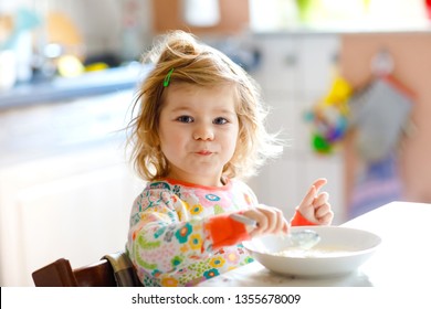 Adorable Toddler Girl Eating Healthy Porrige From Spoon For Breakfast. Cute Happy Baby Child In Colorful Pajamas Sitting In Kitchen And Learning Using Spoon.