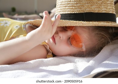 Adorable Toddler Girl Laying On Sun Bed And Hiding From Sun Under Straw Hat. Happy Summer Time Childhood Concept.
