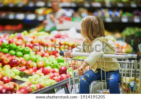 Adorable toddler girl sitting in the shopping cart in a food store or a supermarket. Little kid going shopping. Healthy food for kids