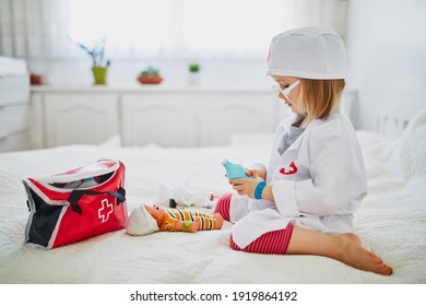 Adorable Toddler Girl In White Coat Playing Doctor And Giving Medical Care To Her Doll. Children And Role Games.