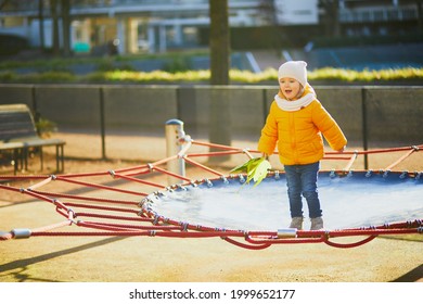 Adorable Toddler Girl In Yellow Jacket Having Fun On A Trampoline On A Fall Or Spring Day. Little Kid On A Playground. Outdoor Activities For Kids