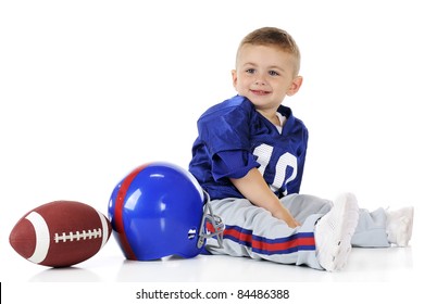 An Adorable Toddler In His Football Uniform.  Beside Him Sits His Helmet And Ball.  Isolated On White.