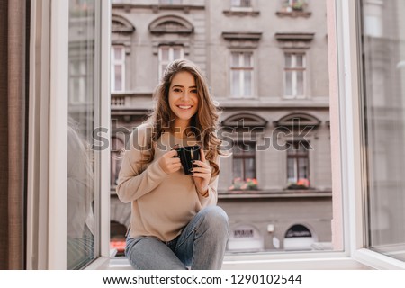 Adorable woman expressing positive emotions while posing on sill with cup of latte. Indoor photo of stunning female model with dark hair wears jeans and sitting beside window.