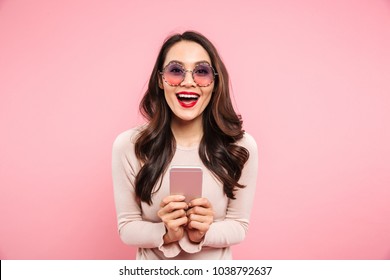 Adorable Woman With Red Lips And Beautiful Brown Hair Chatting Or Using Internet On Cell Phone Over Pink Background