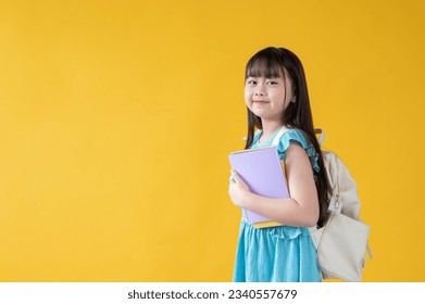 An adorable young Asian girl with books in her hand and a backpack is standing against an isolated yellow studio background with copy space. kindergarten, kids, children, elementary school student - Shutterstock ID 2340557679