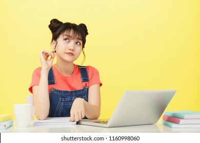 Adorable Young Asian Woman Looking Up In Thoughts While Studying With Laptop And Notepad At Desk On Yellow Background 