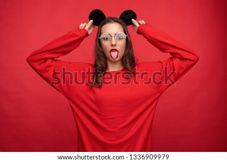 Adorable young brunette in red sweatshirt and black mouse ears posing on red backdrop and showing tongue at camera 