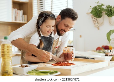 Adorable young daughter with lovelu father in aprons applying kutchup on fresh kneaded homemade dough for pizza. - Powered by Shutterstock