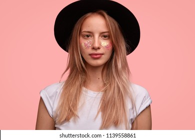 Adorable young European female model with glitter on face, wears elegant black hat, white t shirt, poses over pink background, ready for festival with friends. People, elegance and beauty concept