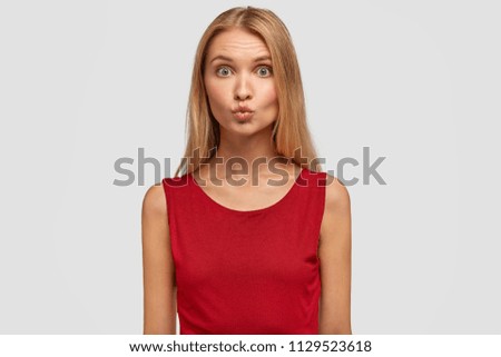 Adorable young female keeps lips rounded, going to kiss her boyfriend, opens eyes widely, dressed in red fashionable clothes, isolated over white background. People and facial expressions concept