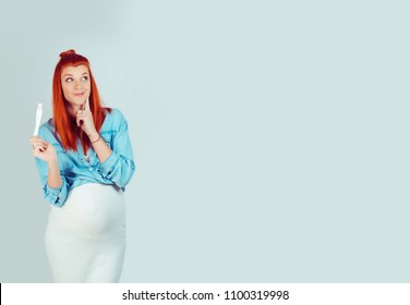 Adorable young girl holding pregnancy test and looking away coquettishly and smiling in dreams isolated on light blue. Thinking about pregnancy, ideas and thoughts. Mixed race model hispanic irish wom