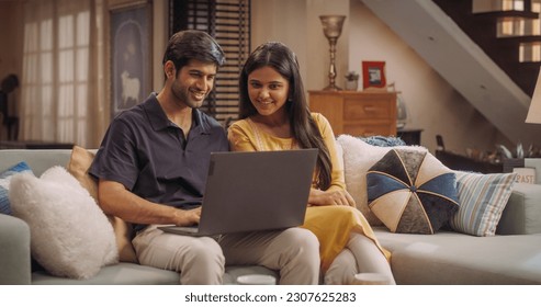 Adorable Young Indian Couple Using Laptop, Sitting on Home Sofa. Exploring Online Content, Discussing Trend Ideas and Making Purchase Decisions Together. Online Shopping Concept. - Powered by Shutterstock