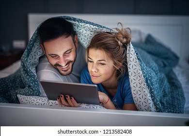 Adorable young relaxed couple lying under blanket on their bad and reading news on tablet.Taking a break for the weekend