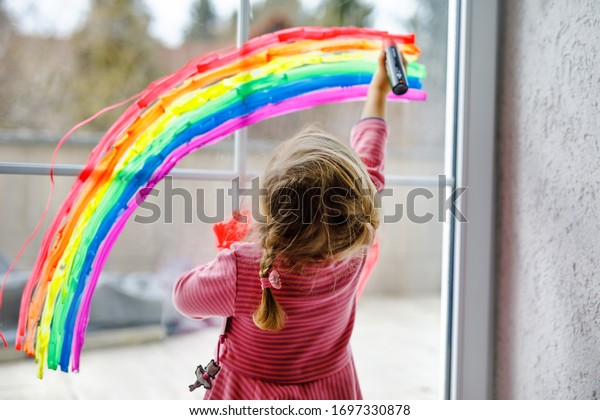 Adoralbe little toddler girl with rainbow painted\
with colorful window color during pandemic coronavirus quarantine.\
Child painting rainbows around the world with the words Let\'s all\
be well.