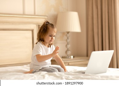 Adorible Little Girl Sitting On Bed At Home, Looking At Laptop Screen Waving Hands Make Distance Video Call. Video Call. Online Communication.