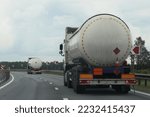 ADR hazardous goods transportation on road. White semi trucks liquid gas tanker with copy space place blank on barrel drive on asphalt motorway at summer day, Back view. 