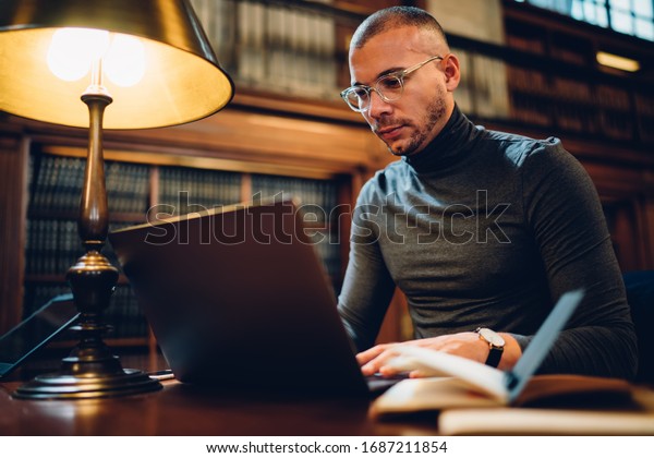 Adult 30 years old male student spending time in\
university library for learning, intelligent man in optical bifocal\
spectacles searching scientific information via app on modern\
laptop computer