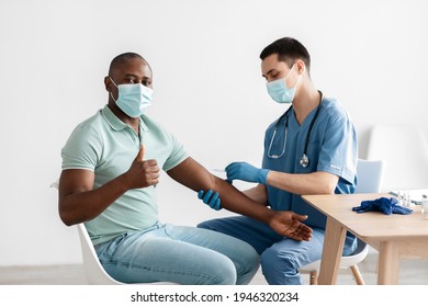 Adult African American Guy Approving Procedure, Gets Covid Vaccine In Clinic, Male Nurse Injecting Into Shoulder. Young Doctor In Protective Mask Makes Shot Of Patient, Guy Show Thumbs Up, Empty Space