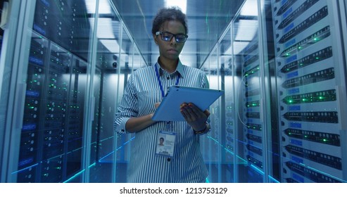 Adult African American woman using tablet while walking among server racks in data center corridor and doing diagnostics