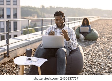 Adult African male wearing denim shirt and headset holding online conference over portable computer on rooftop, female colleague working on blurred background. - Powered by Shutterstock