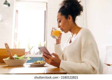 Adult African Woman Looking On Her Phone And Drinking Orange Juice While Eating Avocado Toast And Salad In Cozy Kitchen At Home