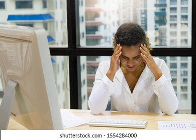 Adult American black woman sitting at computer desk using two hands touching her head because of stress due to overwork in the office surrounded by glass of window and have high-rise building views.