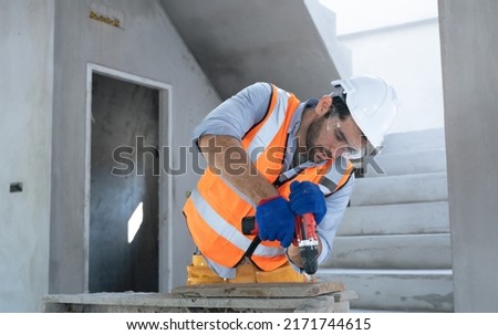 Adult American Hispanic male engineer builder renovating with drill in mask helmet and glove.Handsome Technician using cordless drill in safety first concept.House building under construction site.