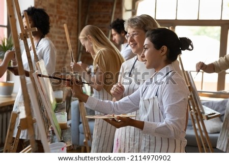 Adult art school Indian student girl in craft apron drawing in paints at easel, holding palette, paintbrush, taking help, advice of mature teacher. Class of artists enjoying creativity in studio