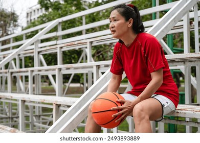 Adult Asian female basketball player holding ball sitting and resting at outdoor stand - Powered by Shutterstock