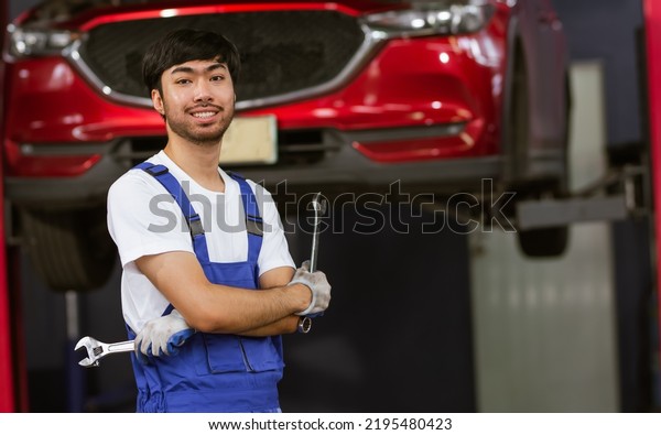 Adult Asian handsome male mechanic wearing
uniform, crossed arms, posing with confidence, standing in garage
at car or automobile maintenance service center or shop with copy
space. Industry Concept