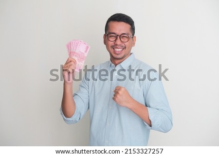 Adult Asian man clenched fist while holding paper money and showing happy expression