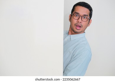 Adult Asian man hiding behind blank white billboard. Peeking out with shocked expression